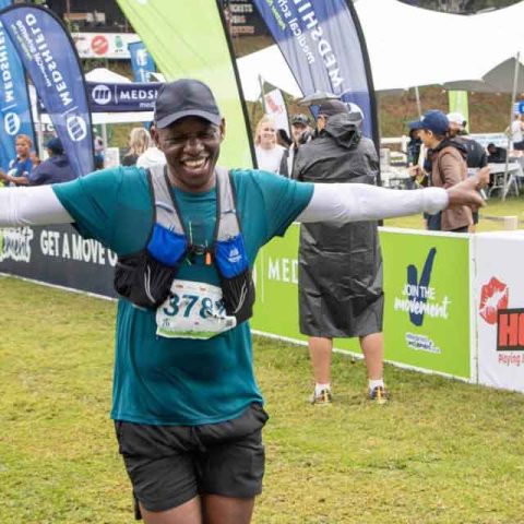A smiling runner with arms outstretched crosses the finish line at Marks Park during an outdoor race event, wearing a teal shirt and race bib 3783. Event banners, staff, and fellow participants fill the background.