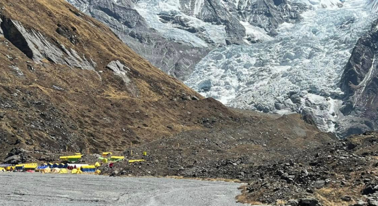 A lone hiker with a backpack walks toward a base camp, marked by a cluster of yellow tents at the foot of snow-covered mountains under a clear blue sky. Rocky terrain and sparse grass cover the ground.