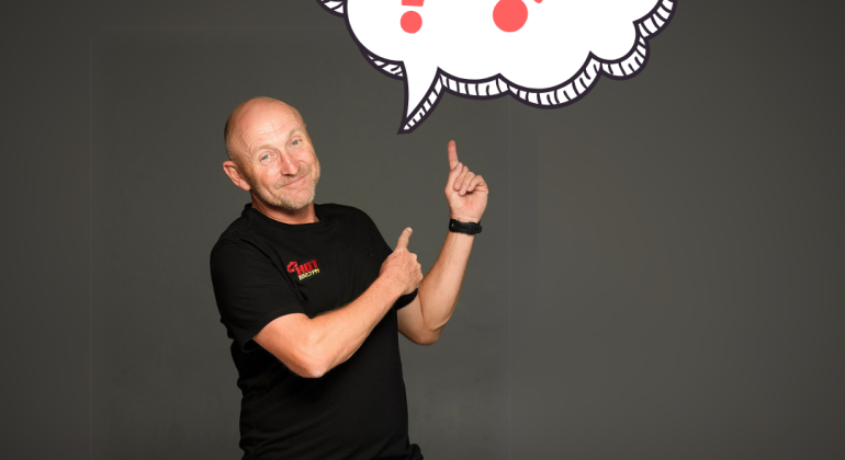 A middle-aged man in a black shirt and jeans stands, smiling and pointing upward at a large illustrated thought bubble filled with red question marks on a dark background.