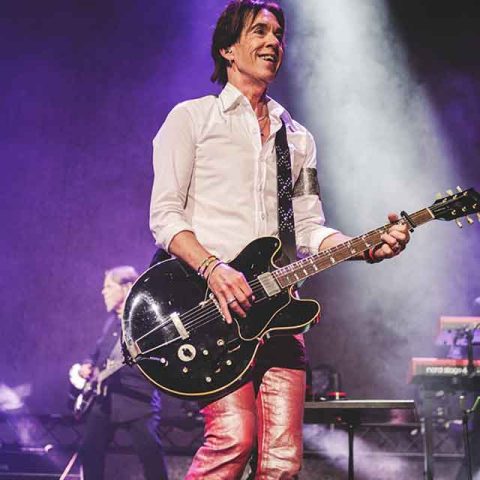 A musician in a white shirt and pink pants plays a black electric guitar on stage, smiling under purple lights, with another Roxette band member and keyboards visible in the background.