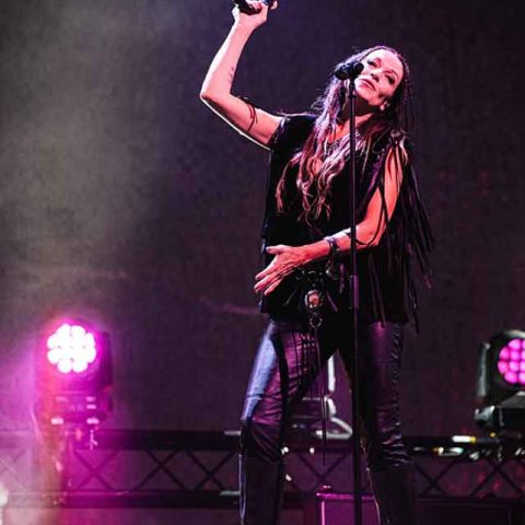 A female musician, reminiscent of Roxette band members, performs on stage in black leather pants and a fringed top, holding a tambourine above her head and singing into a microphone, with vibrant stage lights shining behind her.