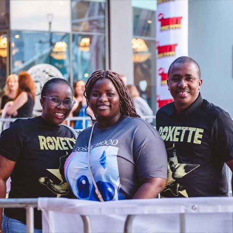 Three people smiling and posing together outdoors—two in matching black ROXETTE shirts, possibly fans of the Roxette band members, and one in a grey T-shirt with a blue graphic. Other people and lights are blurred in the background.