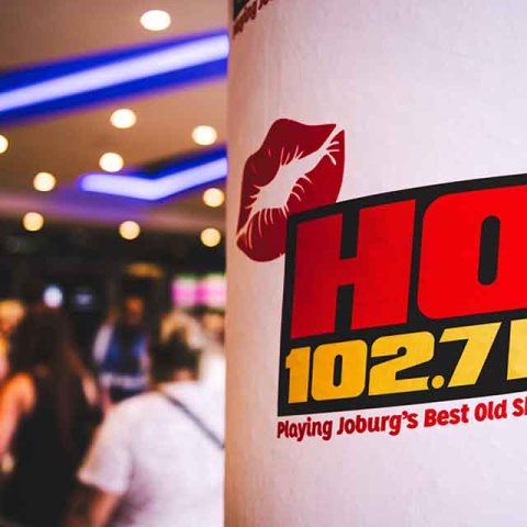 A poster with a red lipstick kiss mark and HOT 102.7 FM in bold letters is displayed on a pillar; blurred people and bright lights create a lively scene, reminiscent of the energy at a Roxette band members concert.
