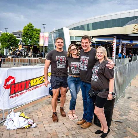 Four people stand smiling together outside Sun Bet Arena, wearing matching dark event t-shirts like Roxette band members. Metal barricades and a HOT 102.7 FM banner are visible, with a cloudy sky overhead.