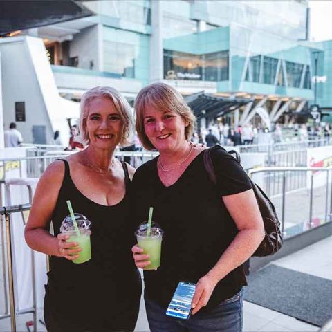 Two women stand outdoors in front of metal barriers, holding green drinks with straws. Both are smiling and casually dressed, reminiscent of Roxette band members. Modern glass buildings and a crowd are visible in the background.