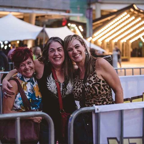 Three women stand together smiling with arms around each other at an outdoor event near metal barriers—channeling the camaraderie of Roxette band members. People and tents appear in the background, along with string lights overhead.