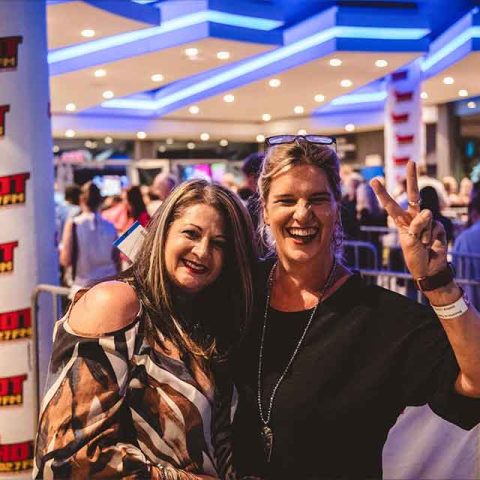 Two women smile at the camera in a lively, brightly lit event space. One flashes a peace sign while people queue under blue lights near a Red Cliffs sign and Hot FM banners, reminiscent of excited fans waiting to see Roxette band members.