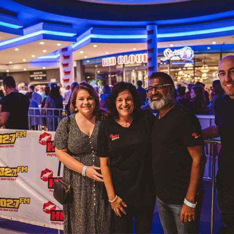 Four people, resembling Roxette band members, stand smiling in front of a crowd and a Hot 102.7 FM banner at an indoor event with blue lighting. A Red Cloud Spur restaurant sign is visible in the background.