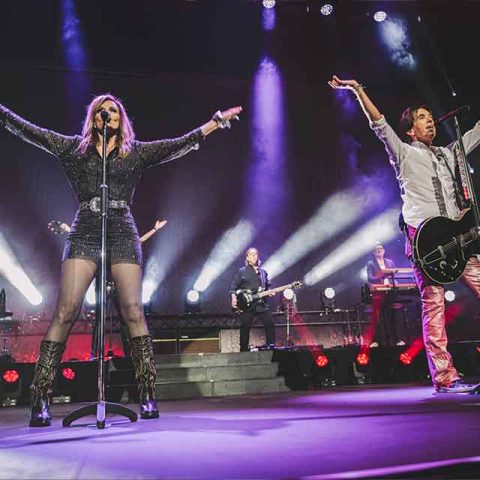 A band performs on stage with dramatic lighting. Two Roxette band members—a female singer and a male guitarist—stand at the front with arms raised, while other musicians play instruments in the background.