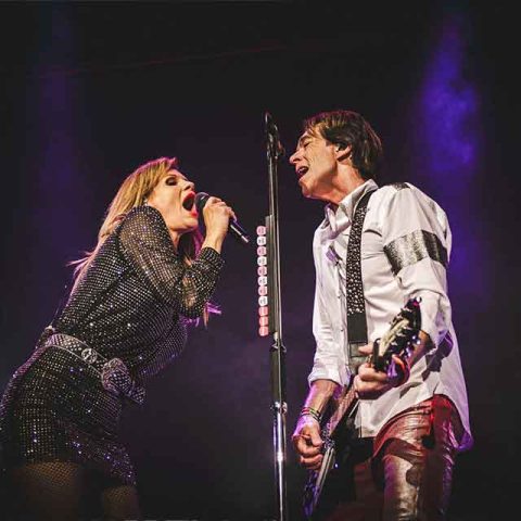 A woman sings passionately into a microphone while a man next to her, resembling one of the Roxette band members, plays guitar and sings. Both are on stage under dramatic lighting, with a dark background and purple stage lights behind them.
