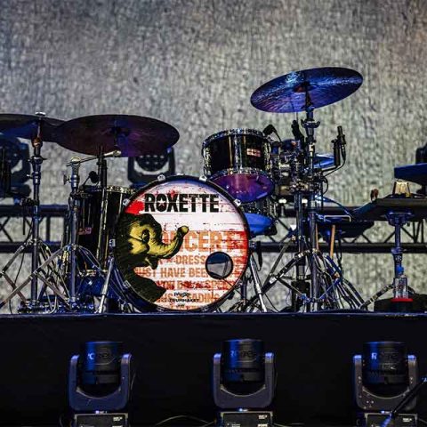 A drum set on stage with a custom bass drum featuring the word ROKETTE and a yellow lizard image, reminiscent of Roxette band members iconic setups. Stage lights, equipment, and a shimmering silver curtain complete the scene.