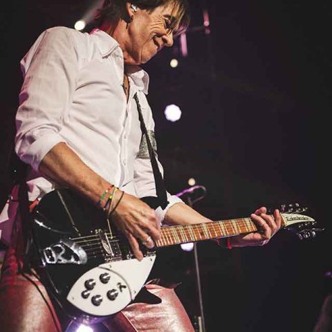 A musician, reminiscent of Roxette band members, wearing a white shirt and shiny pants, plays an electric guitar on stage under bright lights, fully absorbed in the performance.