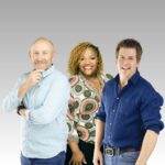 Hot Breakfast team stand smiling in front of a gray gradient background, as if ready to enjoy a Hot Breakfast: a bald man in a light blue shirt on the left, a woman with blonde braids and a patterned shirt in the center, and a man in navy blue on the right.
