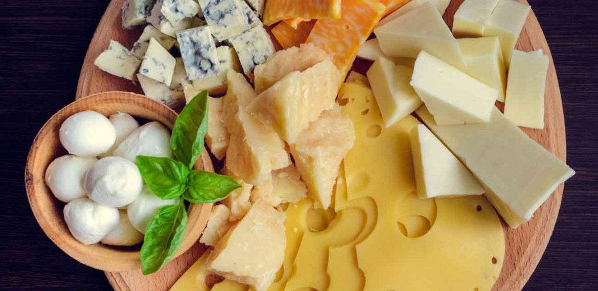 A round wooden board with assorted cheeses, including Swiss, mozzarella balls, blue cheese, cheddar, colby jack, parmesan wedges, and garnished with fresh basil leaves, on a dark surface.