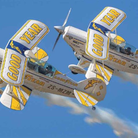 Two white and yellow biplanes with Goodyear branding fly closely together in the sky, leaving white smoke trails behind them against a clear blue background.