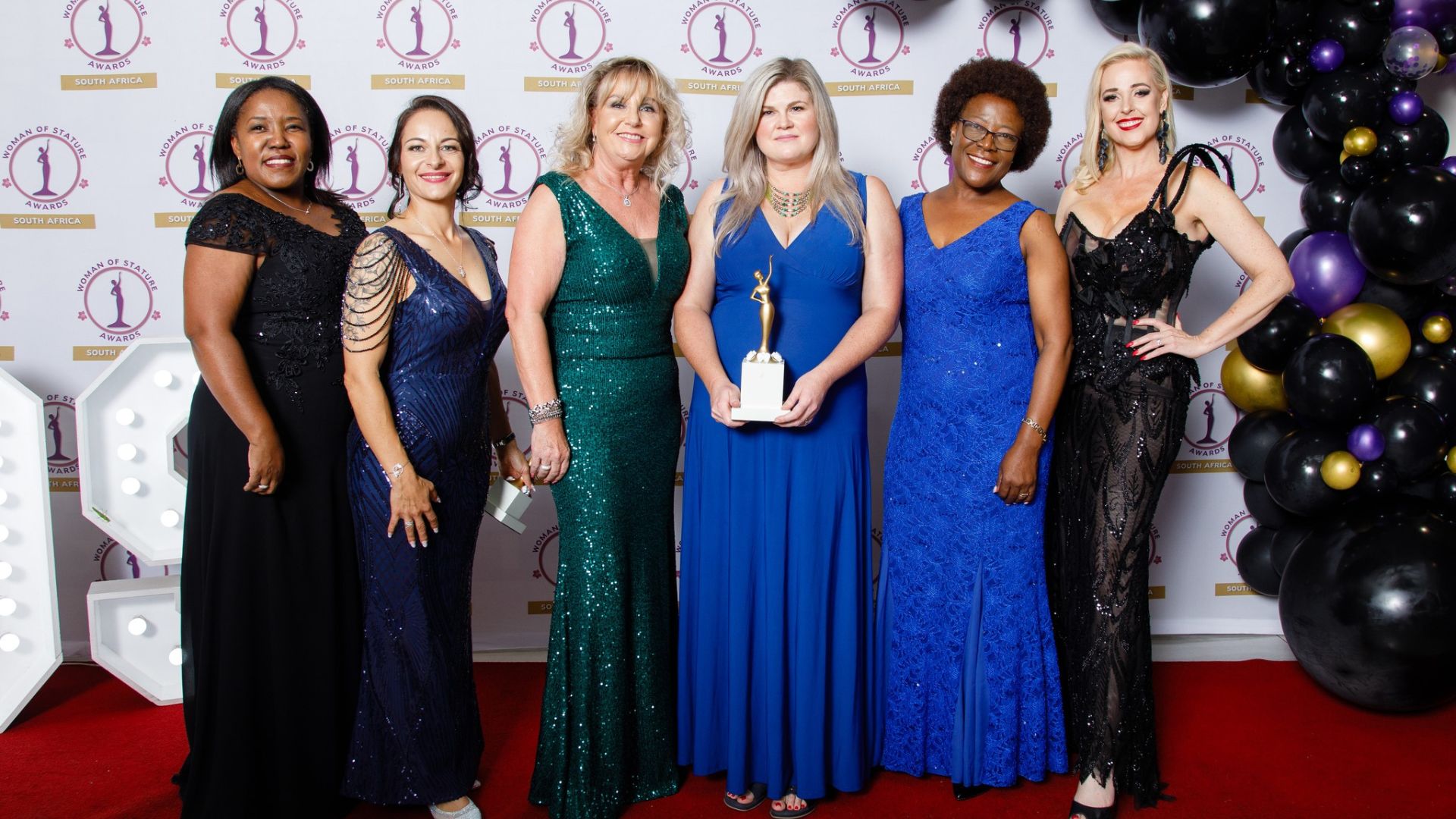 Six women in elegant evening dresses stand on a red carpet at an awards ceremony. The woman in the center holds a trophy, while others smile proudly, with gold and black balloons visible on the right.