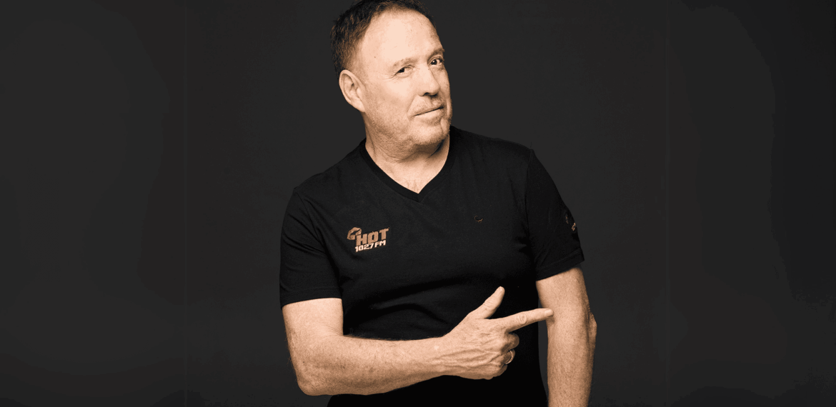 A middle-aged man with short hair wearing a black t-shirt stands against a dark background, smiling slightly and pointing to his left with his right hand.