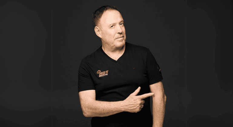 A middle-aged man with short hair wearing a black t-shirt stands against a dark background, smiling slightly and pointing to his left with his right hand.