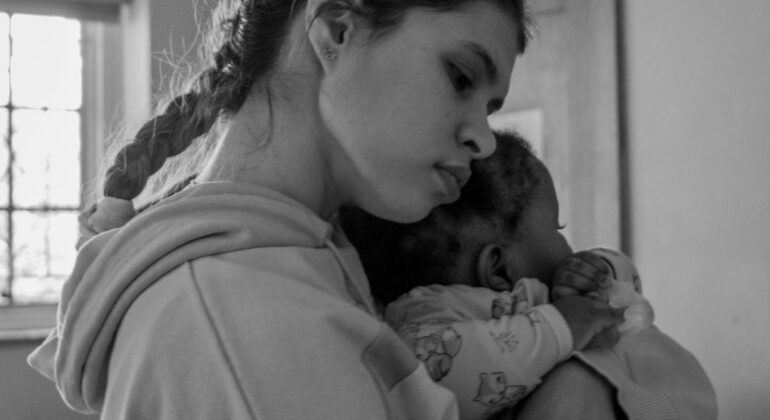 A young woman with a braid hugs and comforts a small baby dressed in patterned pajamas. Soft light enters through a window, giving the scene a calm, caring atmosphere—perfectly reflecting the spirit of Child Protection Week.