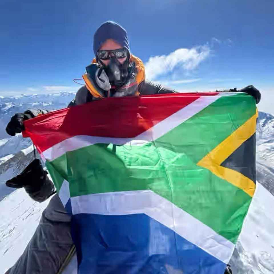 A person wearing cold-weather gear and an oxygen mask holds the South African flag at a snowy mountain summit with a clear sky and distant peaks in the background.