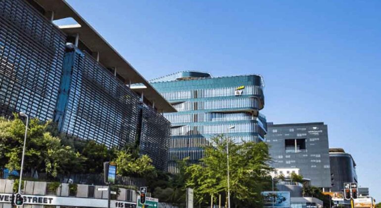A city street with modern glass and steel office buildings, green trees along the sidewalk, pedestrians crossing, and clear blue sky above. Construction barriers are visible near the intersection.