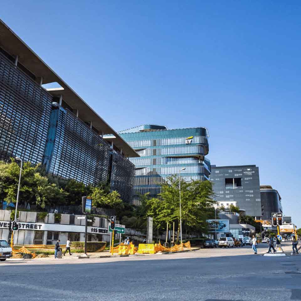 A city street with modern glass and steel office buildings, green trees along the sidewalk, pedestrians crossing, and clear blue sky above. Construction barriers are visible near the intersection.