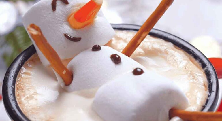 A mug of hot chocolate topped with foam, decorated with a snowman made of three marshmallows, pretzel stick arms and legs, a candy corn nose, and chocolate details for eyes and a smile.