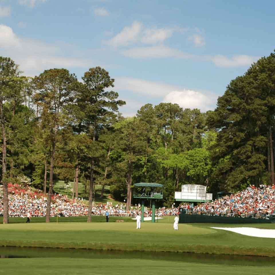 A large crowd watches a golf tournament on a green course surrounded by tall trees, under a partly cloudy sky. Several people stand on the putting green near the flagstick and a sand bunker.