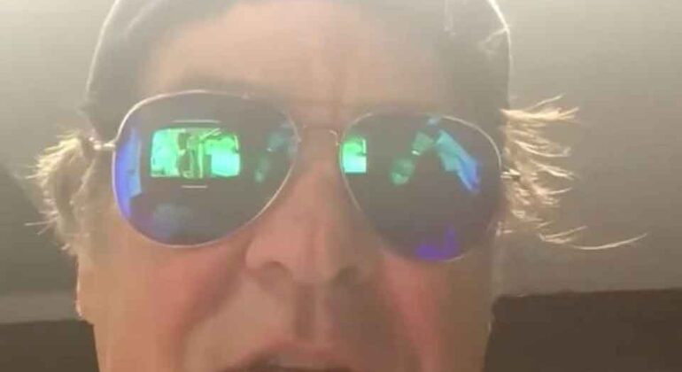 A man wearing a cap and reflective sunglasses looks at the camera with a confused expression. Red question marks surround his head. The background is dim, with bright light above his head.