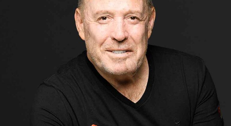 A smiling middle-aged man with short hair and a trimmed beard wears a black t-shirt with a HOT 102.7 FM logo, posing against a plain, dark background.