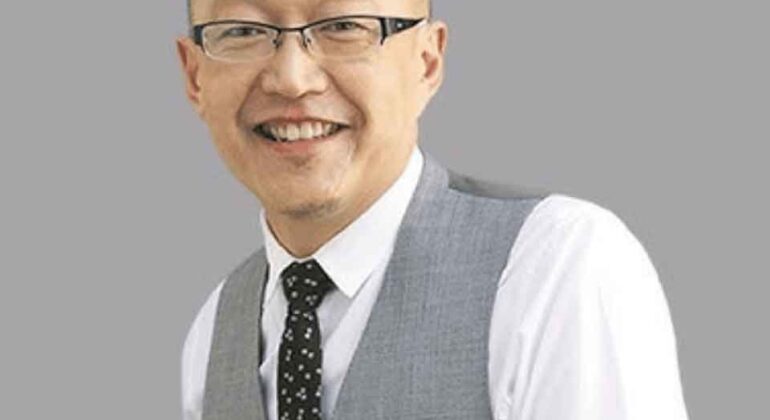 A man with glasses, short hair, and a goatee smiles while wearing a white shirt, grey vest, and black patterned tie, standing against a plain grey background.