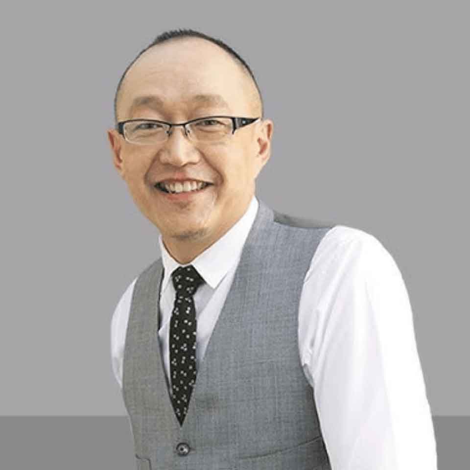 A man with glasses, short hair, and a goatee smiles while wearing a white shirt, grey vest, and black patterned tie, standing against a plain grey background.