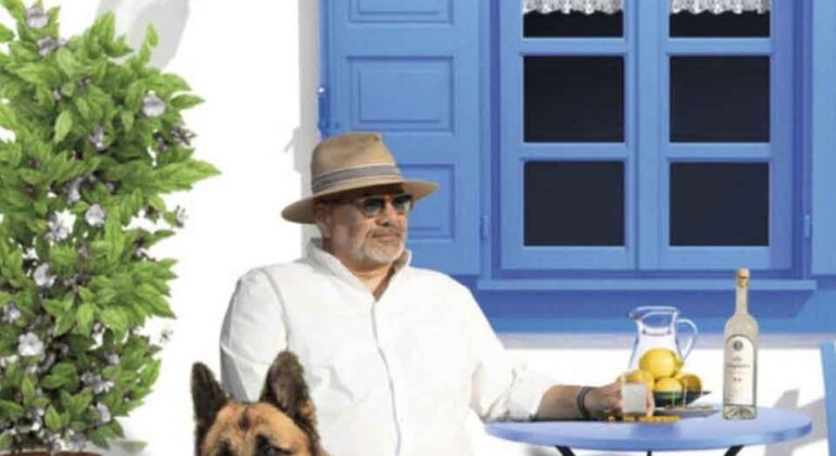 A man in a hat and sunglasses sits at a small blue table with lemonade and a bottle, next to a German Shepherd dog, in front of a white house with blue shutters and a potted plant.