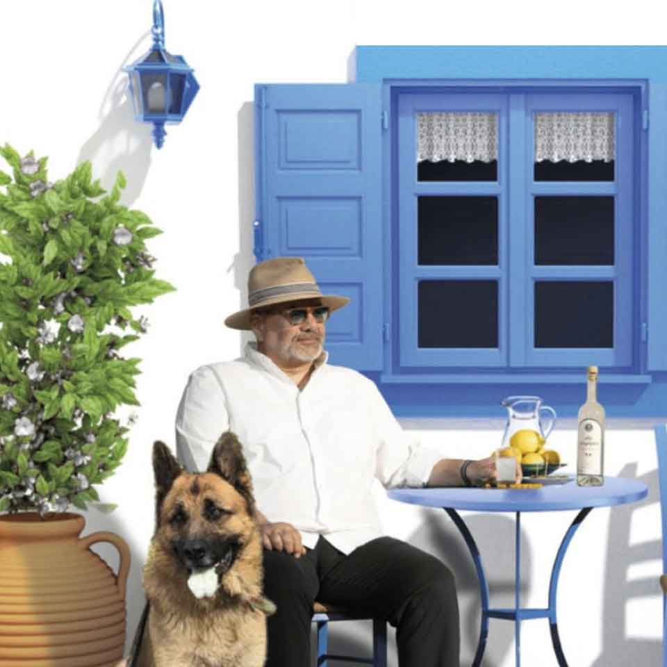 A man in a hat and sunglasses sits at a small blue table with lemonade and a bottle, next to a German Shepherd dog, in front of a white house with blue shutters and a potted plant.