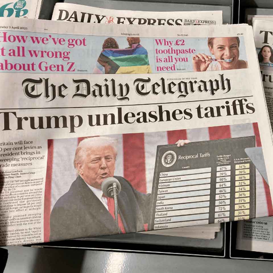 A newspaper front page shows Donald Trump speaking at a podium with a chart of Reciprocal Tariffs beside him. The headline reads, Trump unleashes tariffs. Other newspapers are partially visible underneath.