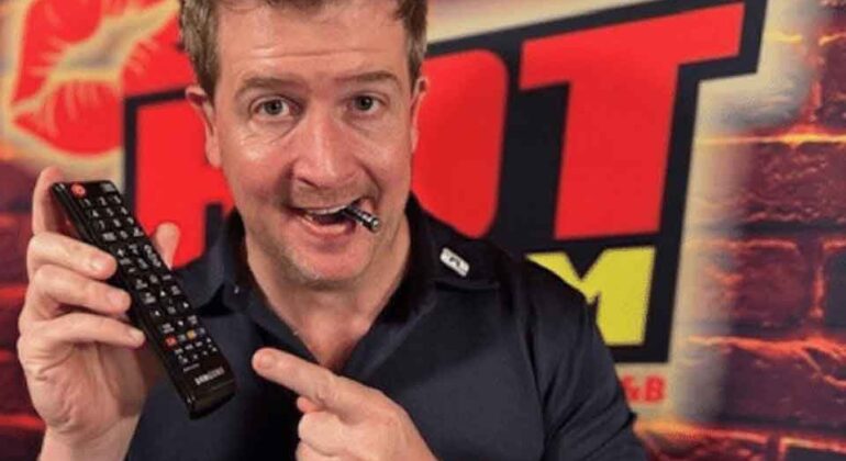 A man in a black shirt smiles while holding a TV remote and pointing at it, with a pen in his mouth. He stands in front of a brick wall with a large HOT FM logo in red and yellow.