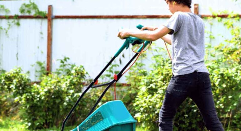 A person wearing a gray t-shirt and dark pants is mowing the lawn with a green lawnmower in a backyard with green grass, bushes, and a white fence.