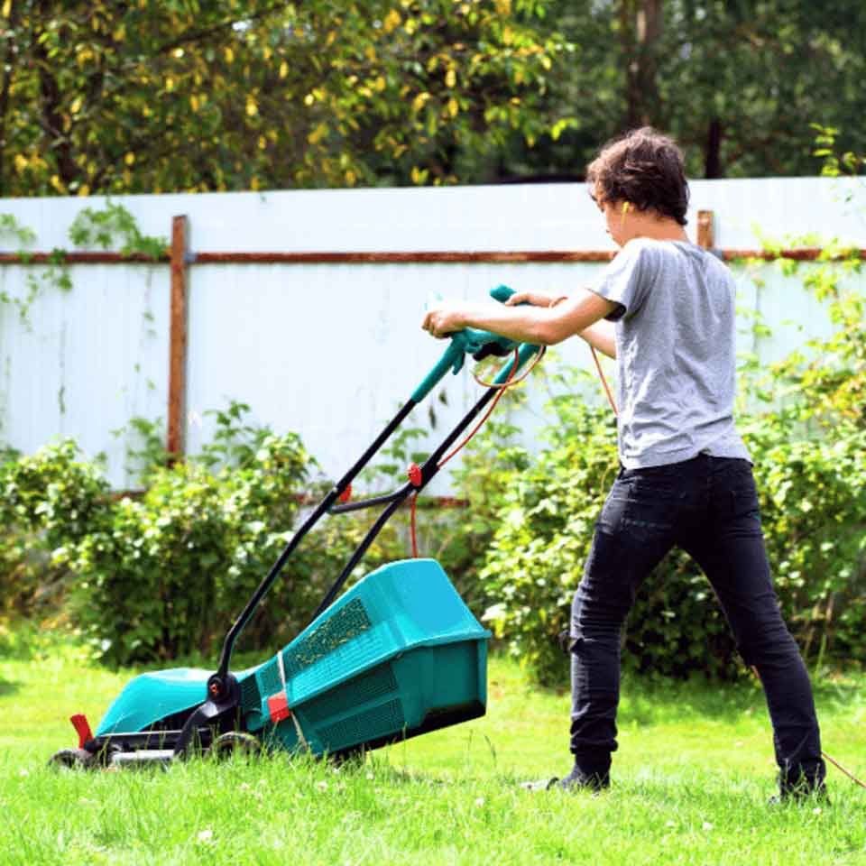 A person wearing a gray t-shirt and dark pants is mowing the lawn with a green lawnmower in a backyard with green grass, bushes, and a white fence.