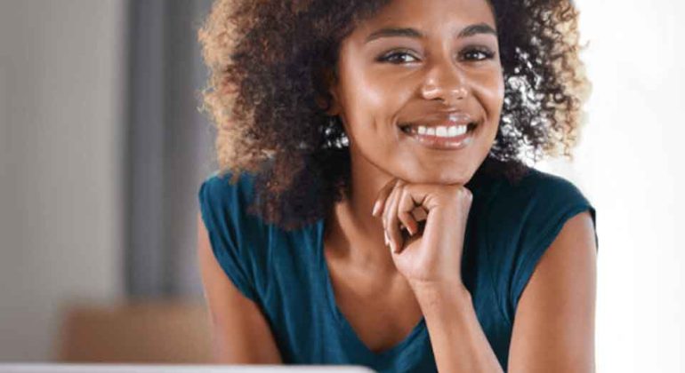 A woman with curly hair wearing a blue shirt smiles while sitting at a desk with a laptop, resting her chin on her hand.