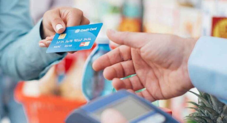 A person hands a blue credit card to another person holding a payment terminal in a grocery store, with a shopping basket filled with groceries visible in the background.