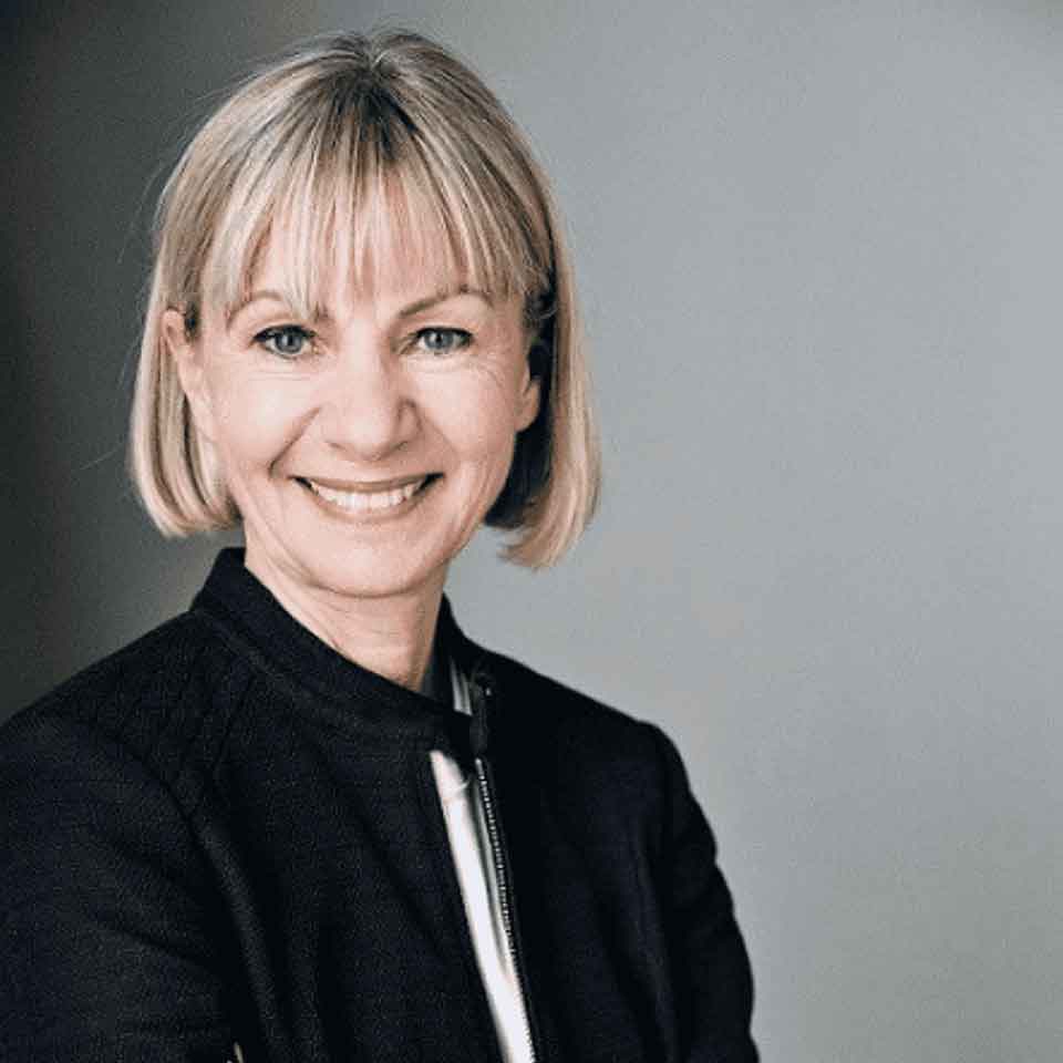 A woman with short blonde hair and bangs is smiling while wearing a dark jacket over a light top, standing against a plain gray background.