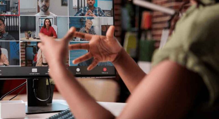 A person wearing headphones gestures while participating in a video conference call with seven other people, shown on a computer monitor in a home office setting.