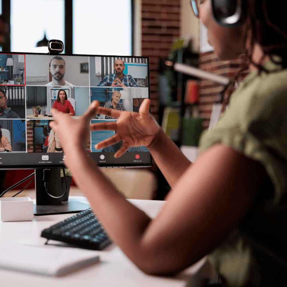 A person wearing headphones gestures while participating in a video conference call with seven other people, shown on a computer monitor in a home office setting.