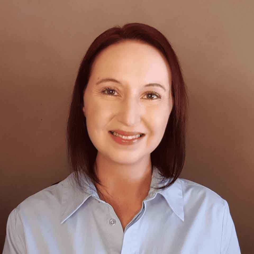 A woman with straight, shoulder-length brown hair smiles at the camera. She is wearing a light blue collared shirt and is posed in front of a plain brown background.