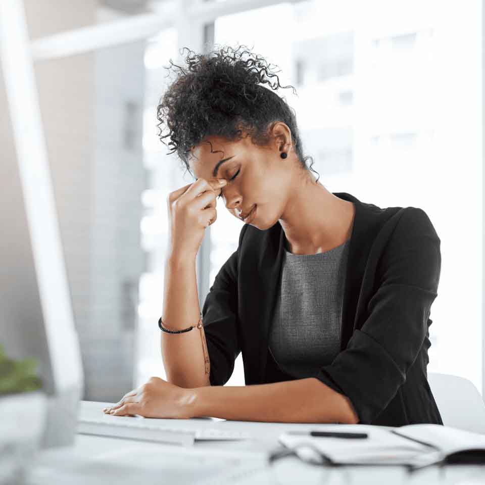 A woman sits at a desk with her eyes closed, pinching the bridge of her nose, appearing stressed or tired. She is dressed in business attire and has a computer and notebook in front of her.