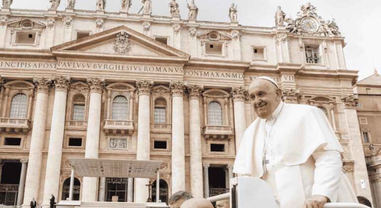 A man in white religious robes and skullcap rides in a popemobile in front of St. Peter’s Basilica, accompanied by security personnel, with ornate architecture and statues visible in the background.
