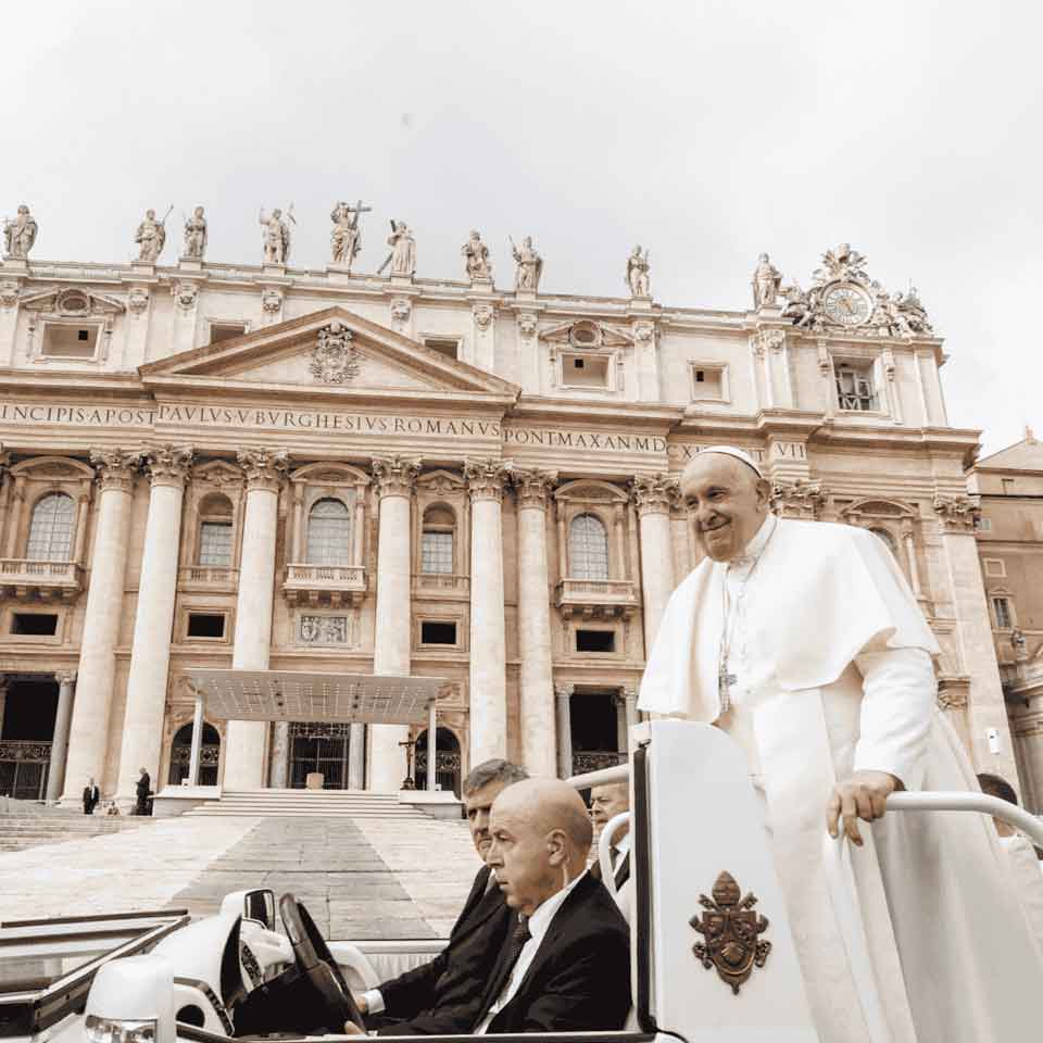 A man in white religious robes and skullcap rides in a popemobile in front of St. Peter’s Basilica, accompanied by security personnel, with ornate architecture and statues visible in the background.