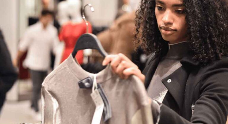 A woman with curly hair examines a silver shirt on a hanger in a clothing store, appearing thoughtful as she shops among racks of clothes.