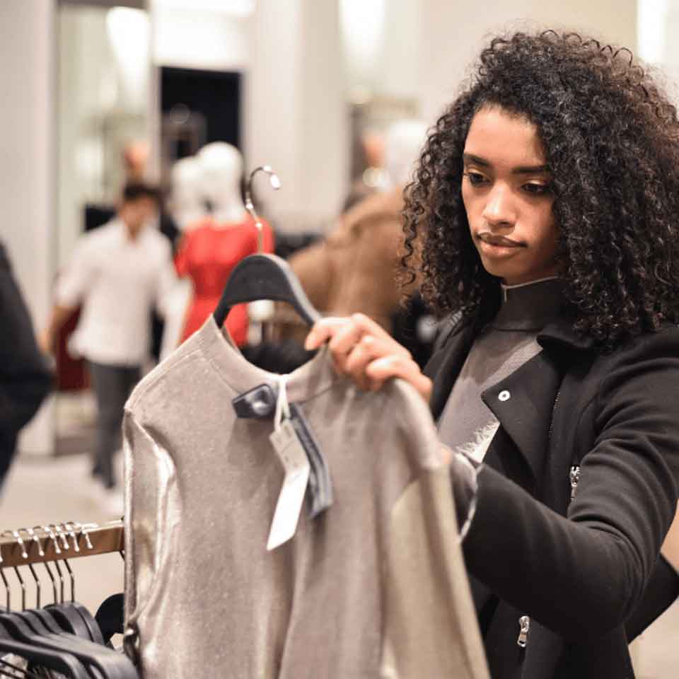 A woman with curly hair examines a silver shirt on a hanger in a clothing store, appearing thoughtful as she shops among racks of clothes.