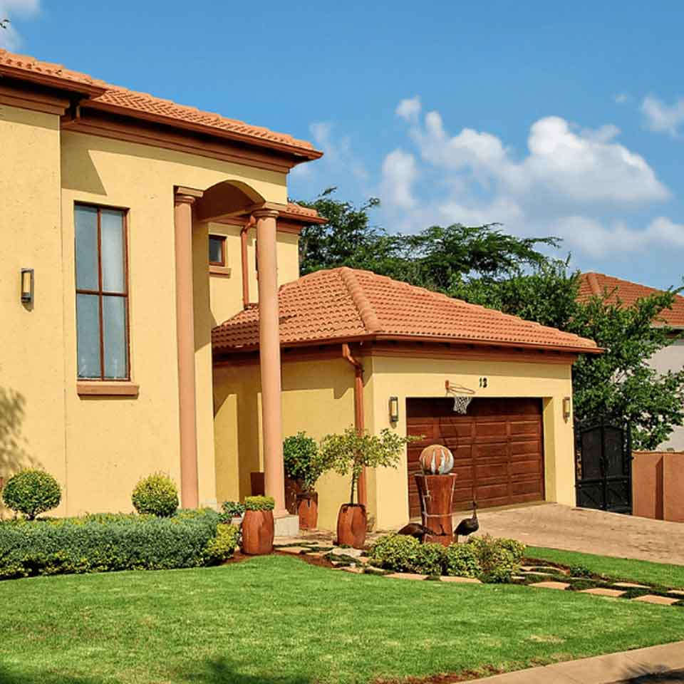 A modern house with a yellow exterior, red-tiled roof, large windows, and a well-maintained lawn with shrubs. A basketball hoop is mounted above the garage door, and the sky is blue with scattered clouds.